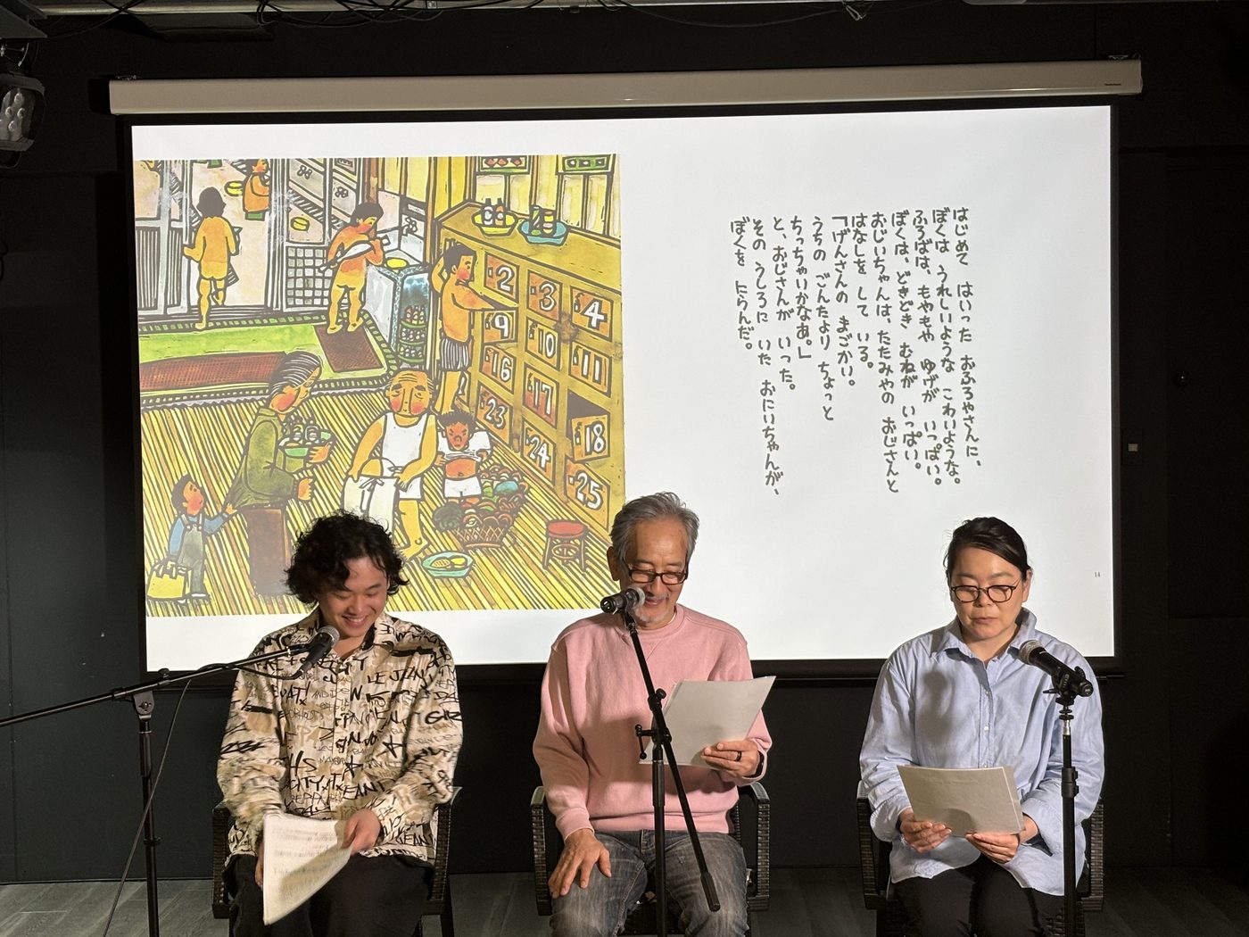 スクリーンに大きく映した絵本を背に、椅子に座って朗読する受講生たち